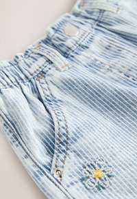 Šviesiai mėlyni džinsiniai šortai su vertikaliomis juostelėmis. Yra kontrastingos siūlės, sidabrinis mygtukas ir mažas geltonos ir baltos gėlės siuvinys.