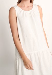 Weißes, ärmelloses Leinenkleid mit rundem Ausschnitt, gerafften Details an den Schultern und gestuftem Saum. Weicher, strukturierter Stoff.