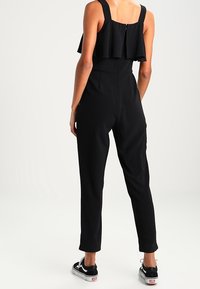 Czarny kombinezon z falbaniastym topem, regulowanymi ramiączkami i dopasowanym krojem. Materiał wydaje się teksturowany. Stylizowany z czarnymi trampkami.