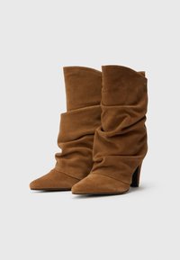 Bruine suède slouchy enkellaarsjes met spitse neuzen en hoge hakken. Heeft zachte plooien en een klein accent vlak bij de enkel.