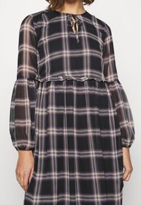 Musta ja ruudullinen mekko, jossa on lyhennetty, läpinäkyvä yläosa. Pitkät hihat joustavilla suutinna, avainaukko-nauhakiinnitys ja röyhelöreunat.