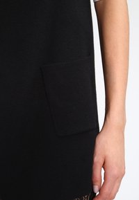 Prenda de punto negra con un bolsillo rectangular en la parte delantera. La textura parece suave. Un borde de encaje es visible en el borde inferior.