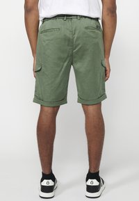 Grüne Baumwollshorts mit umgeschlagenen Säumen, seitlichen Cargo-Taschen und Gesäßtaschen. Getragen mit schwarz-weißen Turnschuhen.