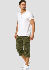 Hvit T-skjorte med korte ermer og tre knapper, kombinert med mørkegrønn kamuflasje cargo-short. Modellen har på seg hvite sneakers og står foran en grå bakgrunn.