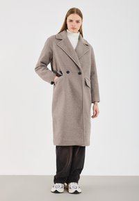 Manteau gris croisé avec une finition texturée, grands revers, deux poches avant, porté sur un pantalon noir et des baskets multicolores.