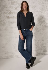 Blusa nera a maniche lunghe con scollatura a V, abbinata a jeans larghi blu con frontale a bottoni e stivaletti neri chunky alla caviglia.