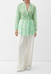 Donna che indossa una blusa a maniche lunghe floreale verde chiaro con vita regolabile e pantaloni bianchi a gamba larga, in piedi contro uno sfondo semplice.