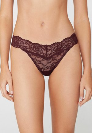 Frau trägt dunkelburgunderrote Spitzen-Bikini-Unterwäsche mit Blumenmuster auf hellem Hintergrund.