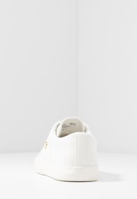 Zapatilla de lona blanca con una superficie texturizada, punta redondeada y una suela de goma suave. Presenta un detalle del logo dorado en el costado.