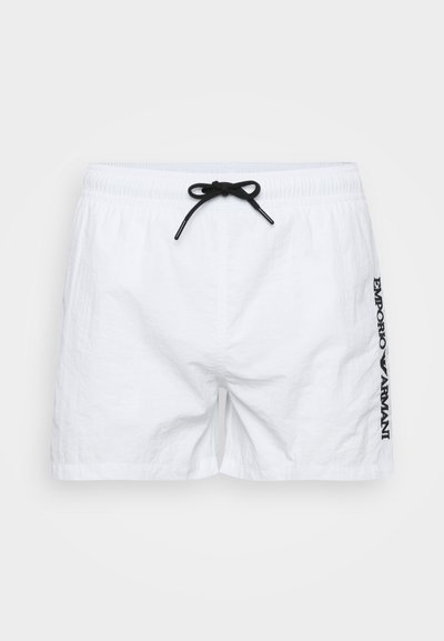 Vita simbyxor i lättviktsmaterial, med elastisk midja och svart dragsnöre samt en svart logotyp tryckt vertikalt på sidan.