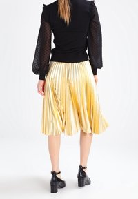 Mujer vistiendo una blusa negra con mangas abullonadas transparentes de puntos y una falda midi plisada metálica dorada, combinada con zapatos negros de tacón cuadrado.