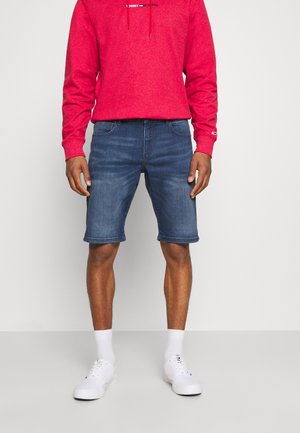 Mand iført en rød hættetrøje, blå denimshorts, hvide sokker og hvide sneakers, stående mod en ensfarvet lys baggrund.