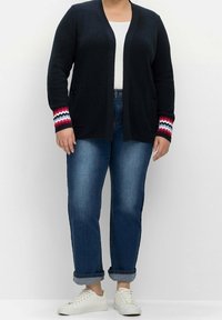Cardigan blu navy scuro con lavorazione a maglia testurizzata, dotato di tasche e dettagli a zigzag multicolore sui polsini. Abbinato a jeans blu risvoltati e sneakers bianche.