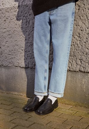 Personne portant un jean bleu clair retroussé, des chaussettes blanches et des chaussures noires en cuir sans lacets, debout sur un pavé en briques à côté d'un mur texturé.