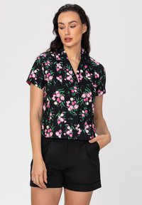 Chemise noire à manches courtes avec un motif floral rose, col à revers et fermeture à boutons. Associée à un short noir ajusté avec des poches.