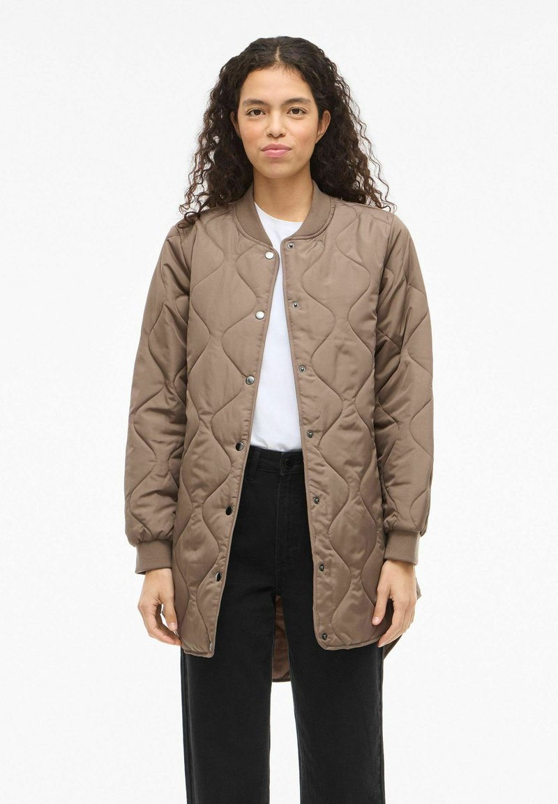 Ruda dygsniuota striukė su spaudėmis, ribotos apdailos rankogaliais ir šiek tiek pratęstu nugaros įkirpimu. Dėvima ant baltos marškinėlių ir juodų kelnių.