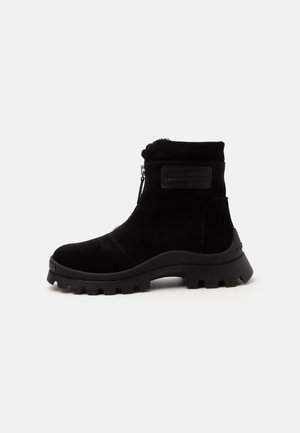 Bottine noire avec semelle épaisse à crampons, fermeture éclair latérale et patch de marque sur la cheville extérieure, en matériau semblable au daim.