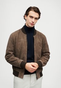 Young man wearing a brown bomber jacket over a black turtleneck sweater and light gray pants, standing with hands clasped.