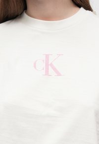 Balta kokvilnas T-krekls ar priekšpusē izšūtu rozā "cK" logo. Dizainā ir iekļauts klasiskā kakla izgriezuma modelis un īsas piedurknes.