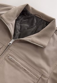 Veste beige zippée avec col côtelé et doublure noire rembourrée, disposant d'une poche poitrine horizontale zippée.