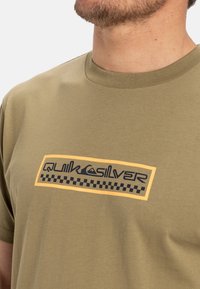 Olivovo zelené tričko vyrobené z bavlny, ktoré má obdĺžnikový čierno-žltý grafický návrh s nápisom "Quiksilver" a vzorom šachovnice.