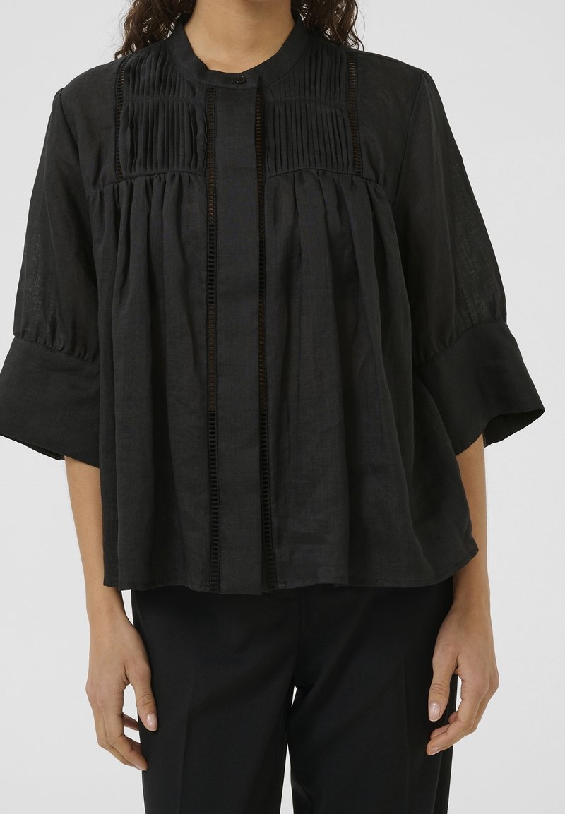 Blouse en lin noir avec un col rond, un corsage plissé et des manches trois-quarts. Présente des coutures verticales et une texture subtile.