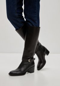 Cizme din piele negre până la genunchi cu toc pătrat, având un accent de cataramă și o textură netedă, purtate cu blugi din denim albastru.