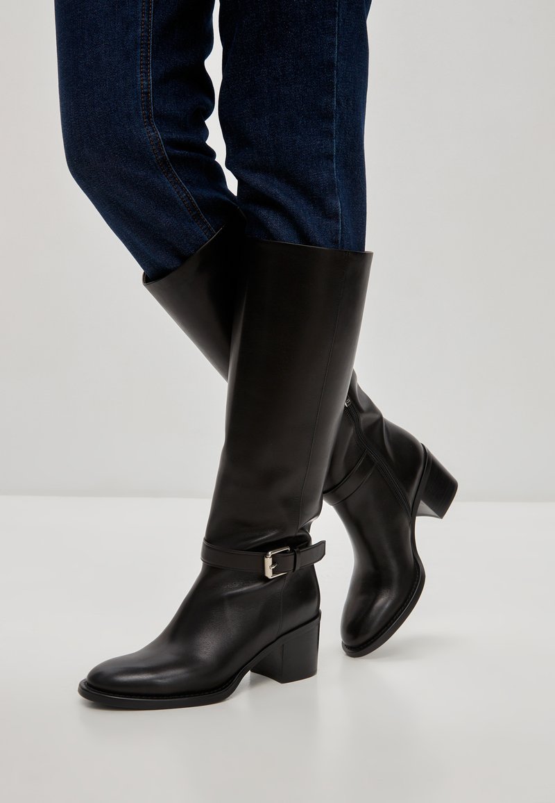 Cizme din piele negre până la genunchi cu toc pătrat, având un accent de cataramă și o textură netedă, purtate cu blugi din denim albastru.