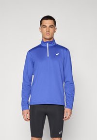 Royal blå langermet treningstopp med glidelås i kragen og hvite detaljer; paret med svarte tettsittende shorts med et logo.
