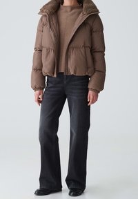Brązowa kurtka puchowa z kołnierzem z futerka ekologicznego, ściągaczami na rękawach i zamkiem błyskawicznym, zestawiona z ciemnymi prostymi jeansami i czarnymi butami.