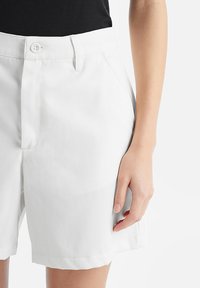 Pantaloni corti bianchi in cotone con taglio dritto, dotati di chiusura con bottone, tasche laterali e una texture liscia. Mano appoggiata sulla vita.