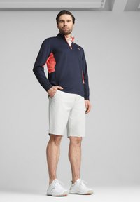 Dunkelblaues Langarm-Polo mit Reißverschluss und korallenfarbenen Akzenten, kombiniert mit weißen Shorts und weißen Sportschuhen. Glattes Material, lässige Passform.