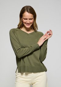 Olivgrön tröja med V-ringning, som har ribbade muddar och nederkant. Tillverkad av mjukt material, med slitsar på sidorna i botten.