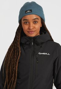 Chaqueta negra con cremallera y un cuello alto, con el logo de O'Neill. Gorro de punto azul con costuras, con una etiqueta tejida, usado sobre cabello largo trenzado.