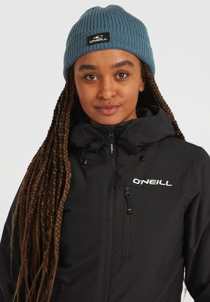 Sort, lynlåsjakke med høj krave og O'Neill-logo. Blå ribbet strikbeanie med et vævet mærke, båret oven på lange, fletninger.