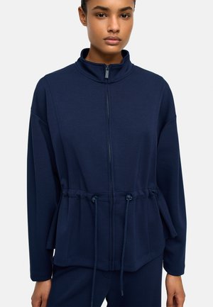 Kobieta w granatowej kurtce zapinanej na zamek z sznurkiem w talii i wysokim kołnierzem, stojąca na jednolitym jasnym tle.