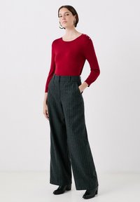 Röd långärmad tröja med knappdetaljer på axeln, kombinerad med högmidjade grå randiga vida byxor och svarta klackstövlar.