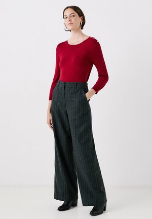 Maglietta rossa a maniche lunghe con dettagli a bottoni sulla spalla, abbinata a pantaloni larghi a righe grigie a vita alta e stivali neri con tacco.