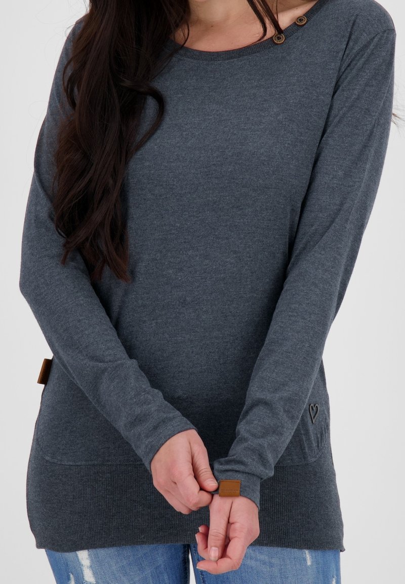 Femme portant une chemise à manches longues gris foncé avec des boutons près de l'encolure et un jean bleu, ajustant le poignet de la manche avec ses mains.