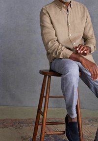 Lichtbruine button-up shirt met een zak, grijze gestructureerde broek en donkergekleurde instapschoenen, zittend op een houten kruk tegen een grijze achtergrond.