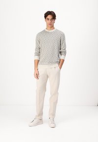 Light gray knitted sweater with diamond pattern, paired with cream-colored trousers and white sneakers, standing against a plain backdrop.