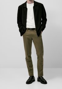 Zwarte jas met ritssluiting over een wit T-shirt, gecombineerd met olijfgroene chinos en zwarte loafers. Eenvoudig ontwerp, met subtiele texturen en kleurcontrasten.