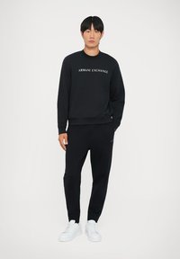 Melns sporta krekls ar "ARMANI EXCHANGE" logotipu, apvienots ar melniem sporta biksēm. Izskatu noslēdz baltas kedas. Gluda auduma, brīvs piegriezums.