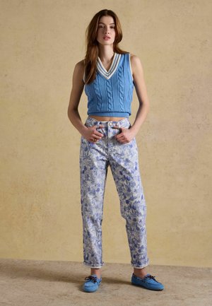 Frau steht mit den Händen in den Taschen, trägt eine blaue ärmellose Strickweste, gemusterte Jeans mit hoher Taille und blaue Mokassins.