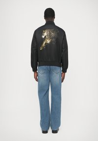 Sort bomberjakke med et gyldent hestemotiv på ryggen, ribbede manchetter og taljebånd, kombineret med lyseblå, vidde cowboybukser.