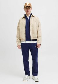 Beżowa kurtka, granatowy sweter, biała koszula, granatowe spodnie i beżowe trampki. Brązowa czapka z emblematem. Slim fit, casualowy design, warstwowy wygląd.