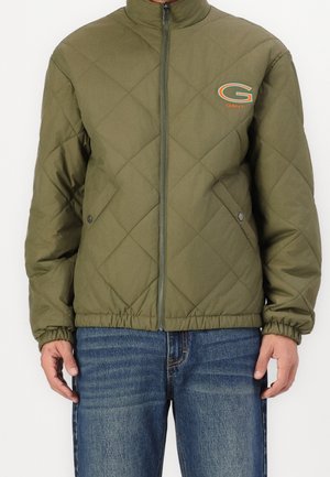 Man draagt een olijfgroene gewatteerde jas met ritssluiting en zakken aan de voorkant, met een oranje en groene "GANT"-logo op de borst, gecombineerd met een blauwe spijkerbroek.