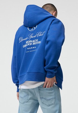 Osoba ubrana w niebieską bluzę z kapturem z napisem „Diverse Sport Club Monaco Grand Hotel” na plecach, w jasnoniebieskich jeansach, zwrócona tyłem.