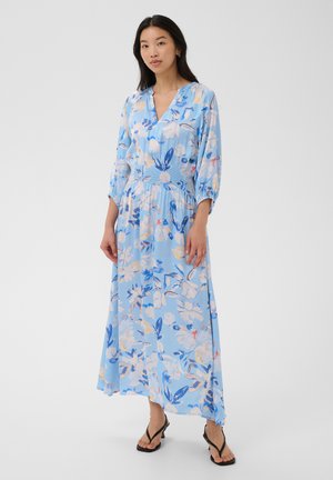 Femme portant une robe longue à manches longues, bleu clair à motif floral, avec une taille cintrée et un col en V, associée à des sandales à talons noires.
