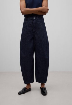 Osoba nosząca ciemne, szerokie, skrócone jeansy, czarne mokasyny wsuwane oraz granatowy bezrękawnik z założonymi skrzyżowanymi rękami.